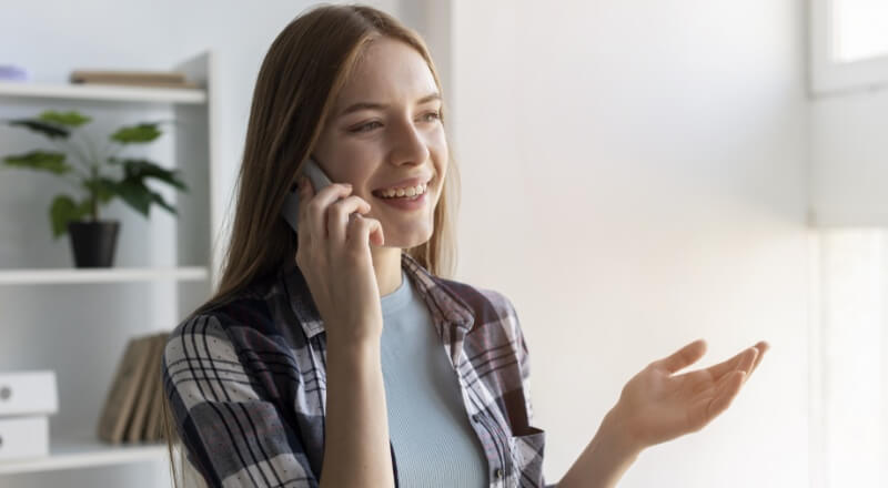 Mulher branca loira sorrindo enquanto aparenta falar com alguém no celular que está enconstado em sua orelha.