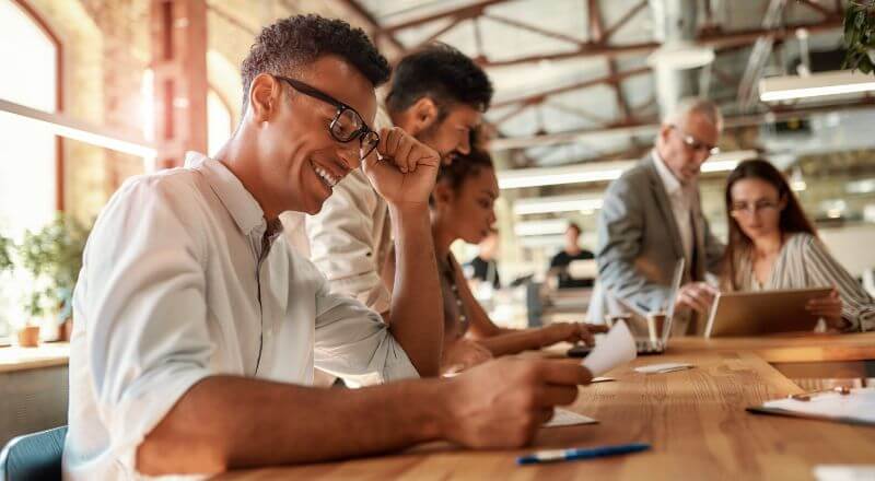 Homem sorrindo para um celular enquanto está sentado em uma mesa com muitas pessoas em volta