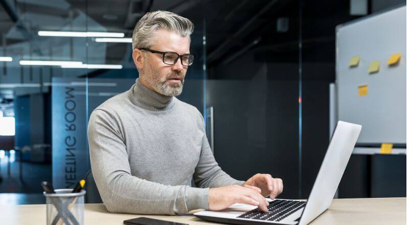 Homem de óculos, com barba e cabelos grisalhos, mexendo em um notebook, num ambiente de trabalho